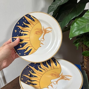 May include: Two round white ceramic plates featuring a gold rim and a celestial design. The graphic depicts a yellow sun face with rays extending into a dark blue background filled with small gold stars. The sun design covers one side of each plate.