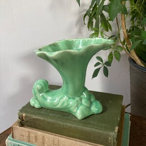 May include: A green ceramic vase with a trumpet-shaped opening and a curled base. The vase is decorated with a textured, leafy design.