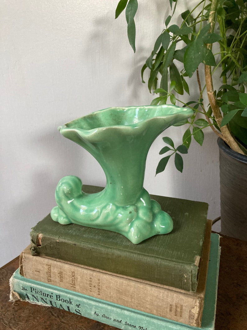 May include: A green ceramic vase with a trumpet-shaped opening and a curled base. The vase is decorated with a textured, leafy design.