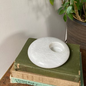 May include: White marble candle holder with a round shape and a hole in the centre. The holder is sitting on top of a stack of three books.