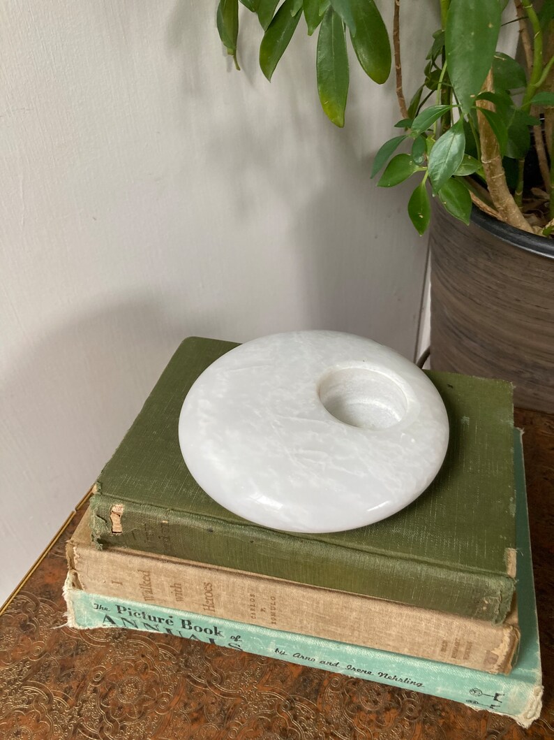 May include: White marble candle holder with a round shape and a hole in the centre. The holder is sitting on top of a stack of three books.