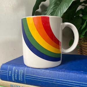 May include: White ceramic mug with a rainbow design. The rainbow arcs across the mug in red, orange, yellow, green, and blue. The mug has a white handle and is sitting on a stack of books.