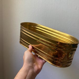 May include: An oval-shaped container with a metallic gold finish and a smooth, reflective surface. The vessel features a series of horizontal ridges running along its exterior walls, creating a textured appearance around the entire perimeter.
