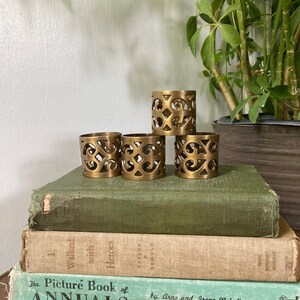 May include: Four gold metal napkin rings with an intricate, cut-out design. The rings are stacked on top of each other on a stack of books.
