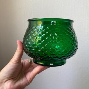 May include: A green glass bowl with a textured, scale-like pattern. The bowl has a rounded shape with a flat base and a wide opening. The bowl is held by a hand.