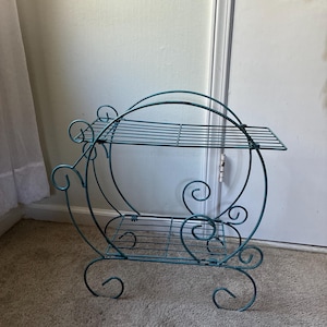 May include: A teal metal plant stand featuring two rectangular wire shelves, a large circular frame, and decorative scrollwork accents. The structure is supported by four curved legs and includes a rounded handle at the top for mobility.