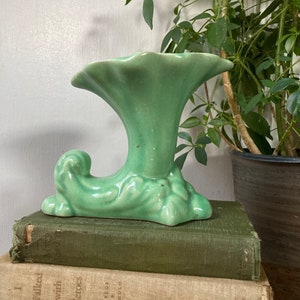 May include: A green ceramic vase with a trumpet-shaped opening and a stylized leaf design. The vase is sitting on a stack of books.