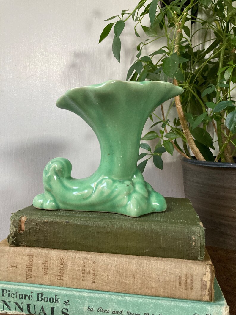 May include: A green ceramic vase with a trumpet-shaped opening and a stylized leaf design. The vase is sitting on a stack of books.