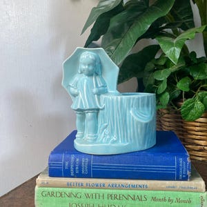 May include: A light blue ceramic planter featuring a young girl holding an umbrella. The planter has a cylindrical base and is set on a stack of vintage books with titles like "The Deepening Stream" and "Gardening with Perennials".