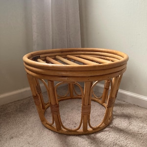 May include: A round, natural-toned rattan side table. The table features a woven top and a cylindrical base with vertical supports and decorative circular accents. The table is approximately 18 inches in diameter and 18 inches tall.