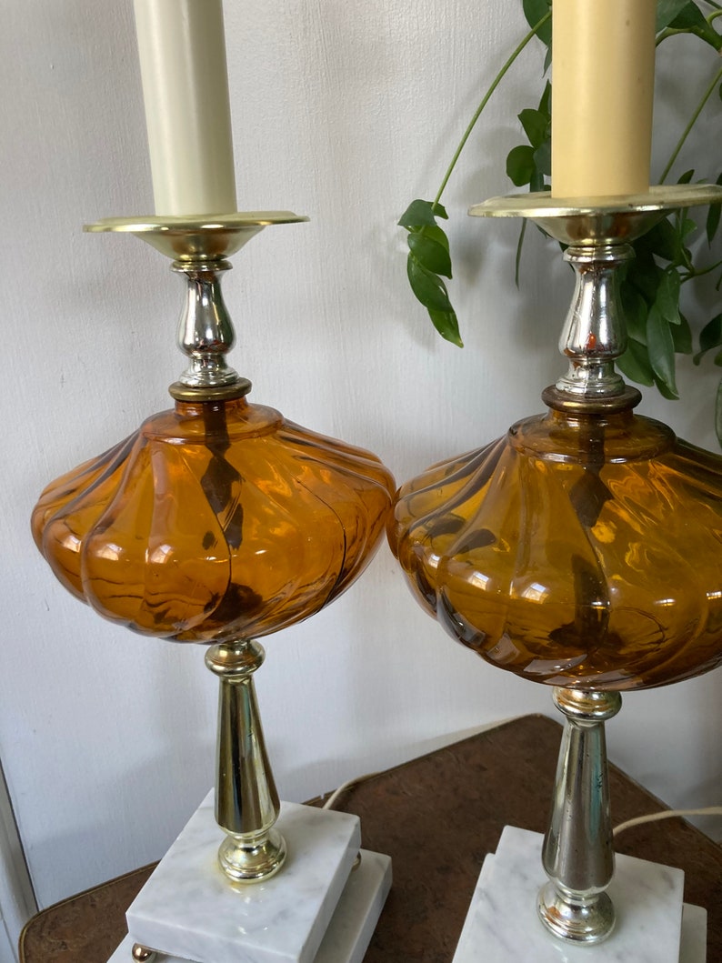 May include: A pair of amber glass table lamps with white marble bases and gold accents. The lamps have a fluted design and are topped with a candle-style bulb.