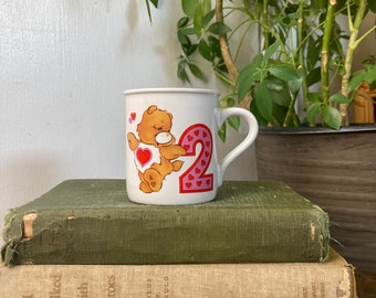 May include: White ceramic mug with a cartoon bear wearing a pink heart and holding a heart-shaped cookie. The mug has a pink number 2 with pink hearts on it.