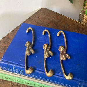 May include: Three brass monkey-shaped hooks. Each hook features a monkey figure at the top and a curved hook at the bottom. The hooks are placed on a blue book with the text "THE OPENING DREAM CROTHY FIELD".