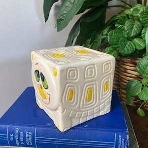 May include: A white ceramic turtle-shaped planter with yellow accents and a painted face. The planter is sitting on a stack of books.