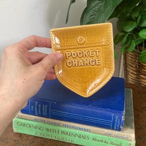May include: A mustard-yellow ceramic pocket change holder with a stitched design and the words "POCKET CHANGE" embossed on the front. The holder is held in front of a stack of books, including one titled "Gardening with Perennials".