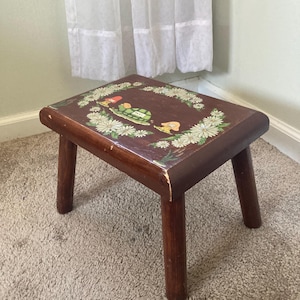 May include: A small, wooden step stool with a dark brown finish. The top is decorated with a floral and whimsical design featuring daisies and cartoon characters. The stool has four sturdy, rounded legs.