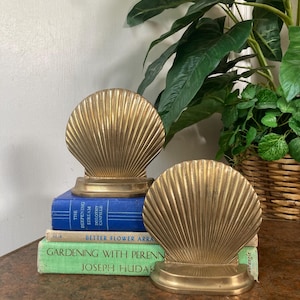 May include: Two gold-colored seashell bookends. The bookends are placed on a stack of books with blue and green covers. The bookends have a detailed, ridged design, and are placed on a wooden table.