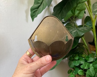 May include: A triangular brass wall pocket with a smooth, metallic finish and a scalloped lower edge. The object features two keyhole-shaped cutouts at the top for mounting purposes, showcasing a weathered patina across its surface.