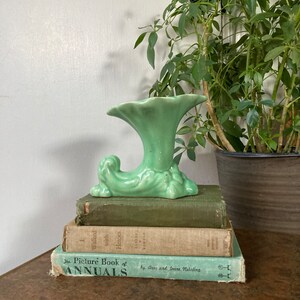 May include: A green ceramic vase with a unique, stylized design. The vase has a wide, trumpet-shaped opening and a base that resembles a curled leaf. The vase is sitting on top of a stack of three books.
