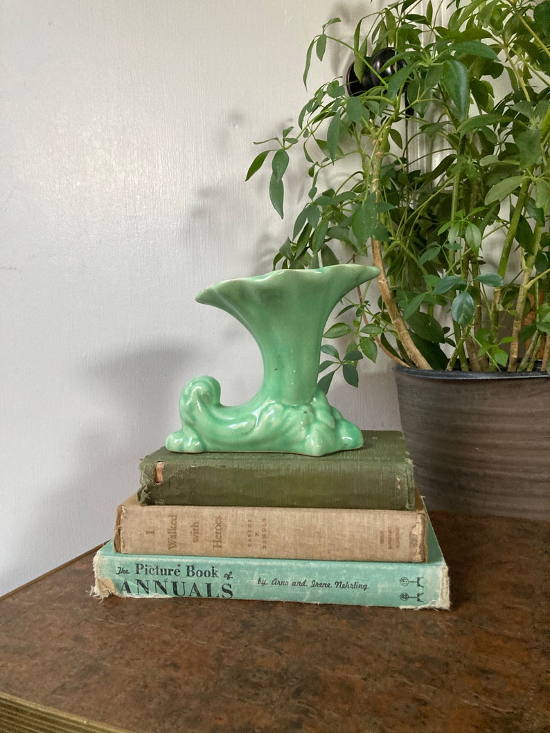 May include: A green ceramic vase with a unique, stylized design. The vase has a wide, trumpet-shaped opening and a base that resembles a curled leaf. The vase is sitting on top of a stack of three books.