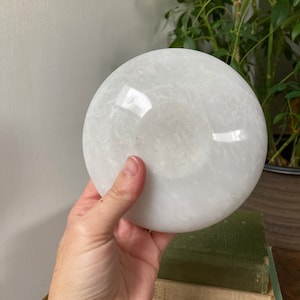 May include: A white selenite disc, also known as a selenite plate, is a smooth, round, and polished piece of selenite crystal. It has a white, opaque appearance with subtle striations and a slightly translucent quality.