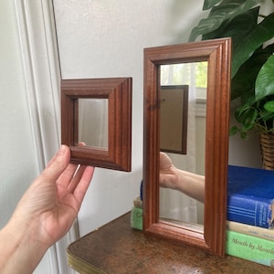 May include: Two wooden framed mirrors. One is square, the other is rectangular. Both have a dark brown finish. The square mirror is held by a hand. The rectangular mirror is standing on a table next to a stack of books.