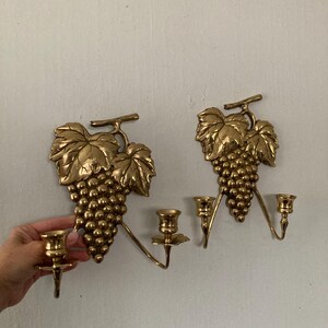 May include: Two gold-colored wall sconces, each shaped like a bunch of grapes with leaves. Each sconce has two candle holders. The sconces are made of metal and are mounted on a light-colored wall.