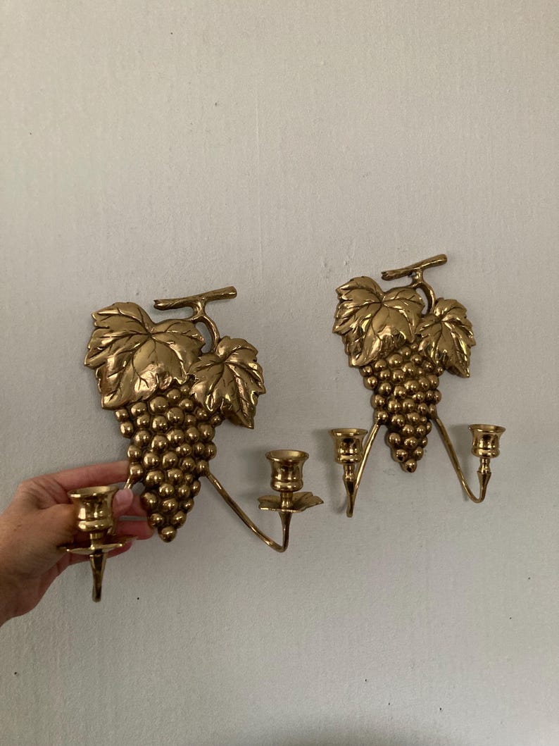 May include: Two gold-colored wall sconces, each shaped like a bunch of grapes with leaves. Each sconce has two candle holders. The sconces are made of metal and are mounted on a light-colored wall.