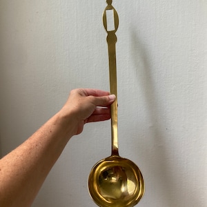 May include: A large, gold-colored brass ladle with a long handle and a decorative top. The handle has a rectangular opening in the center.