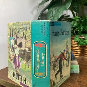 May include: Vintage books titled "Heidi" and "Hans Brinker" are displayed. The books have colorful illustrations and text, including "Companion Library" on the spine. The books are on a table with a gold trim, with greenery in the background.