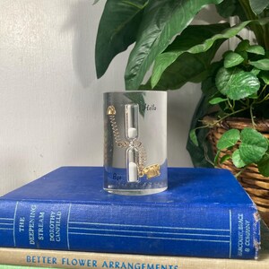 May include: A clear glass hourglass with gold accents and the words "Hello" and "Bye". The hourglass is set on a stack of books with titles like "The Deepening Stream" and "Gardening with Perennials". A green plant is in the background.