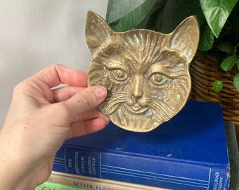 May include: A brass trinket dish shaped like a cat's face. The dish has a distressed finish and is a light gold color.