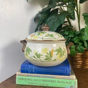 May include: Cream-colored enamelware pot with a lid, adorned with yellow and green floral patterns. The pot has bronze-colored handles and a matching lid finial. It rests on a stack of books with titles like "Better Flower Arrangements" and "Gardening with Perennials."