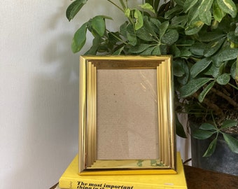 May include: A gold-colored picture frame with a simple, rectangular design. The frame is sitting on top of a stack of three books. The top book has the text "The most important thing in the world" printed on the cover. The second book has the text "Better Flower Arrangements" printed on the cover. The bottom book has the text "Gardening with Perennials Month by Month" printed on the cover.
