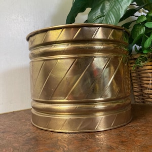 May include: A cylindrical brass planter featuring a polished metallic finish with a repeating diagonal etched pattern. The vessel has two horizontal raised bands encircling the body, one near the top rim and one near the base, providing texture.