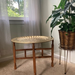 May include: A round brass tray table with a decorative embossed design. The table has a wooden frame with four legs and a cross-shaped support.