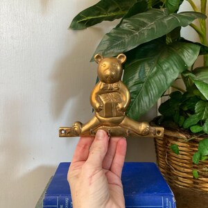 May include: A gold-colored metal bear-shaped wall hook holding a gift. The bear is in a split position with a horizontal bar below with three hooks. The hook is being held in front of a stack of books and a green plant.