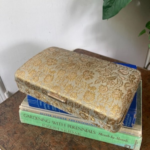 May include: A rectangular, gold-toned box with a floral pattern, resting on a stack of books. The box has a clasp closure and is placed on top of books with titles like "Better Flower Arrangement" and "Gardening with Perennials".