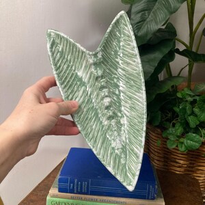 May include: A green ceramic leaf-shaped dish with a white glaze. The dish is textured and has a raised design. The dish is sitting on top of a stack of books.