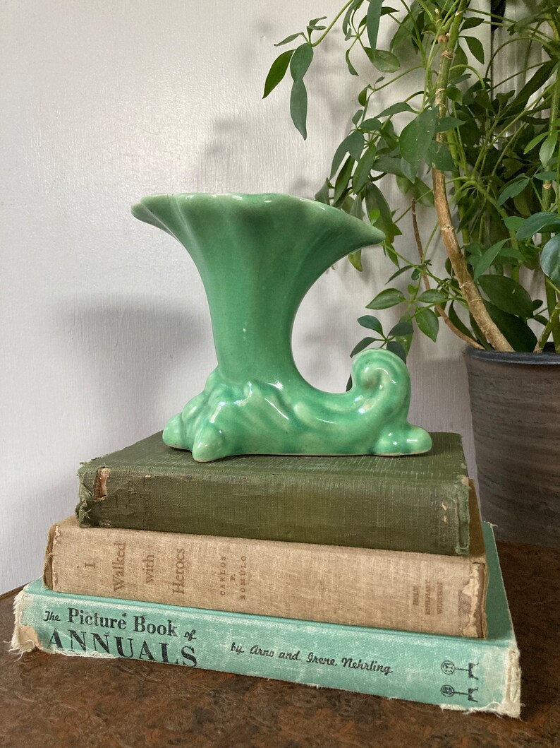 May include: A green ceramic vase with a unique, wave-like design. The vase is sitting on top of a stack of three books.