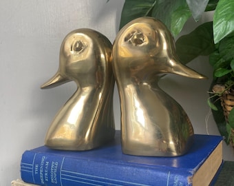 May include: Two brass duck head bookends. The bookends are gold-colored and sit on top of a stack of vintage books with blue and green covers. The bookends are in the shape of a duck's head and neck.