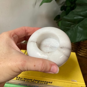 May include: White marble candle holder with a round, slightly indented top. The holder is smooth and has a subtle gray veining.