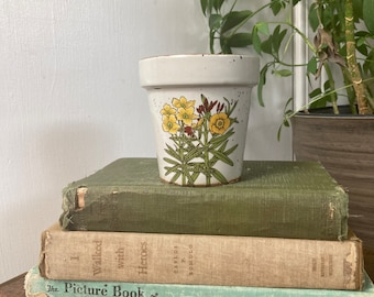 May include: A white ceramic flower pot with a speckled glaze and a floral design featuring yellow flowers and green leaves. The pot is sitting on top of a stack of three books.