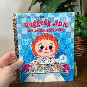 May include: A vintage Little Golden Book titled "Raggedy Ann and the Cookie Snatcher." The cover features a cartoon illustration of Raggedy Ann holding a tray of cookies. The background is blue with a checkered pattern. The book has a gold spine.
