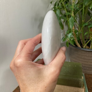 May include: A smooth, white, oval-shaped selenite crystal. The crystal is held in a hand, with the tip of the crystal pointing upwards.