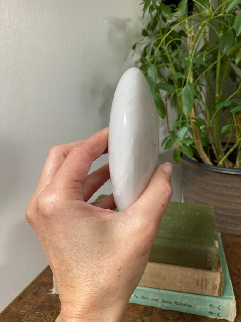 May include: A smooth, white, oval-shaped selenite crystal. The crystal is held in a hand, with the tip of the crystal pointing upwards.