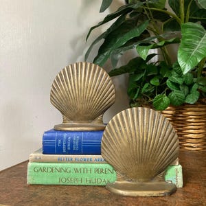 May include: Two gold-colored seashell bookends. The bookends are placed on a table with a stack of books. The books are blue and green. A green plant is in the background.
