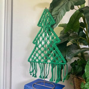 May include: A green macrame Christmas tree decoration. The tree is made of knotted green yarn, with a diamond pattern and fringe at the bottom. It stands on a gold-colored metal stand, and is approximately 10 inches tall.