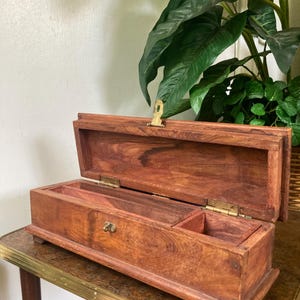 May include: A rectangular wooden box with a hinged lid, featuring a natural brown wood grain finish. The interior is divided into two compartments, and the exterior includes a brass-colored metal latch and hinges for secure closure.