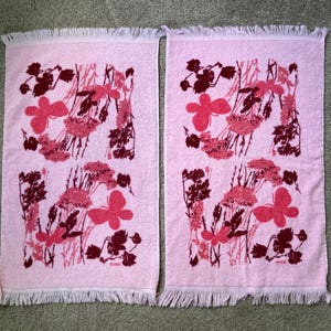 May include: Two matching pink hand towels with a floral design in shades of pink and burgundy. The towels have a fringed edge and a rectangular shape. The floral pattern includes stylized flowers and foliage.